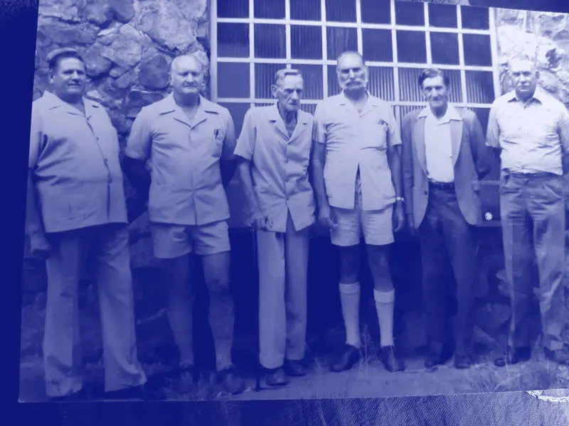 'n Ou afdruk van ses ouer familiemans wat in 'n ry teen 'n klipmuur staan