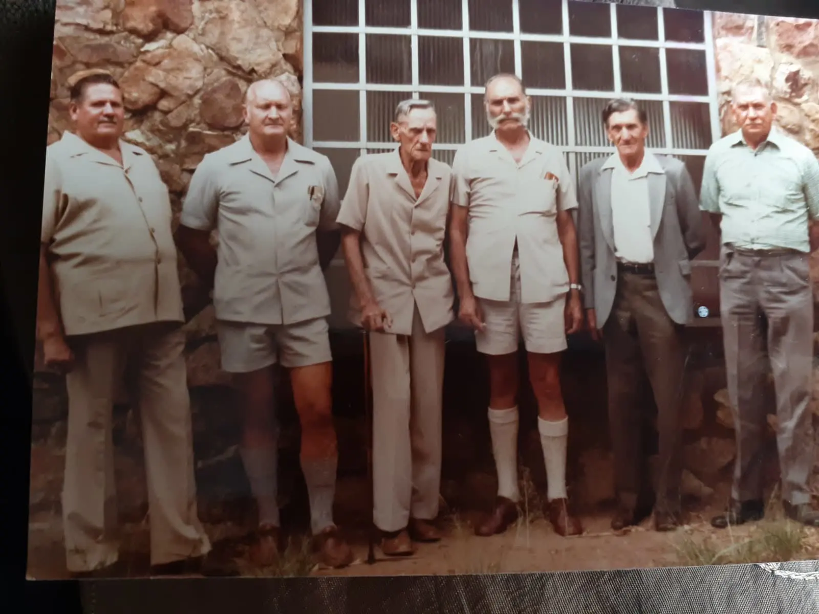 'n Ou afdruk van ses ouer familiemans wat in 'n ry teen 'n klipmuur staan